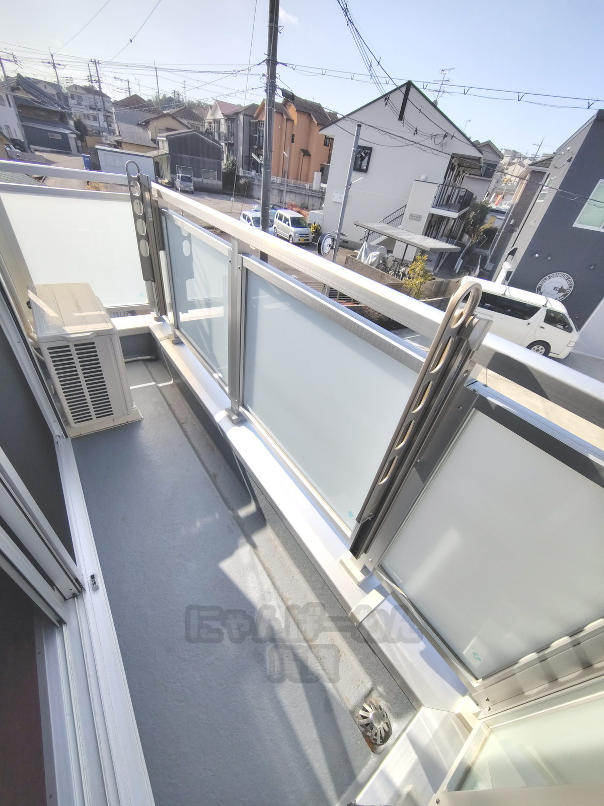 八幡市八幡小松の賃貸物件のバルコニー。日当たりの良い南向き風のベランダ、エアコン室外機、洗濯物干し金具、曇りガラスの手すり、周辺の住宅街の眺望。